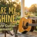 Acoustic guitar resting on a weathered bench on a porch at sunset, with bold yellow text: 'YOU ARE MY SUNSHINE' and smaller subtitle 'Fingerstyle Guitar Arrangement'